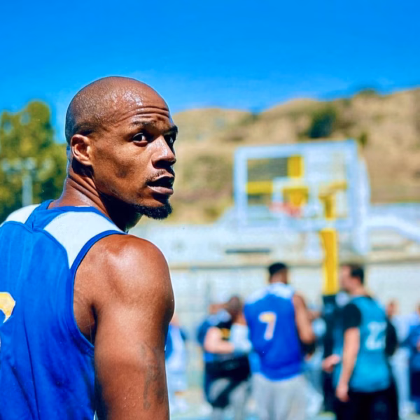 Anthony in a blue basketball jersey on an outdoor court, with other players and a basketball hoop in the background.