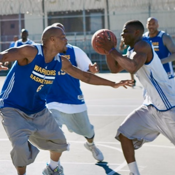 Anthony plays basketball at San Quentin State Prison, wearing a Warriors jersey and dribbling the ball while being guarded by another player.