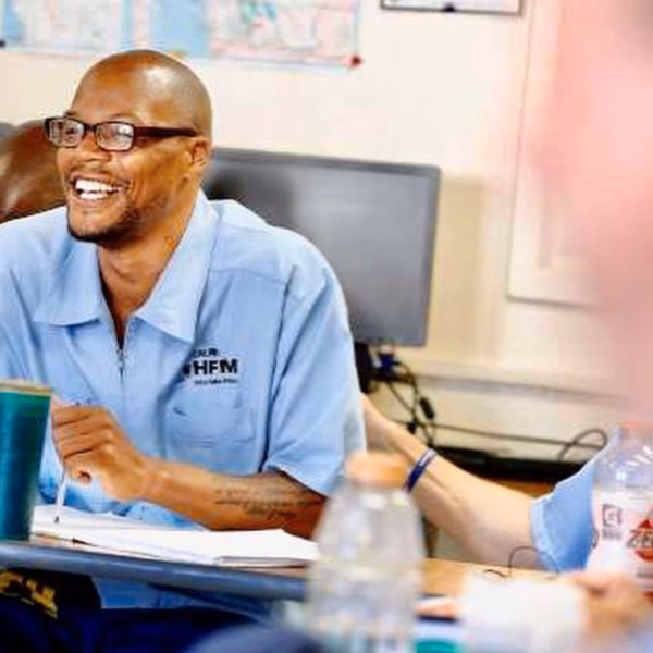 Anthony sitting at a desk in a light blue San Quentin State Prison uniform, smiling while writing.