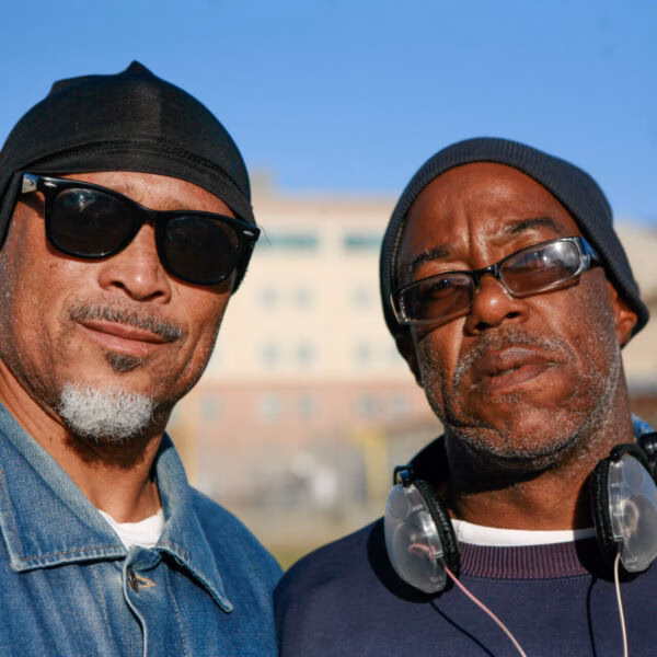 Two men wearing beanies and sunglasses stand side-by-side outdoors against a blurred building.