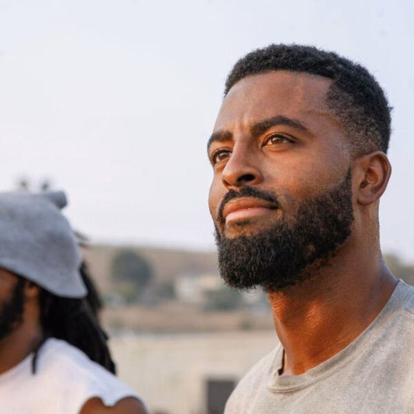 Two men outdoors; a bearded man in a grey shirt looks forward, while another man with dreadlocks and a beanie smiles in the background.