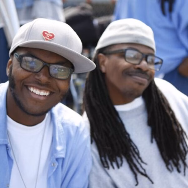 Two men wearing glasses and caps, one smiling broadly, in a casual outdoor setting.