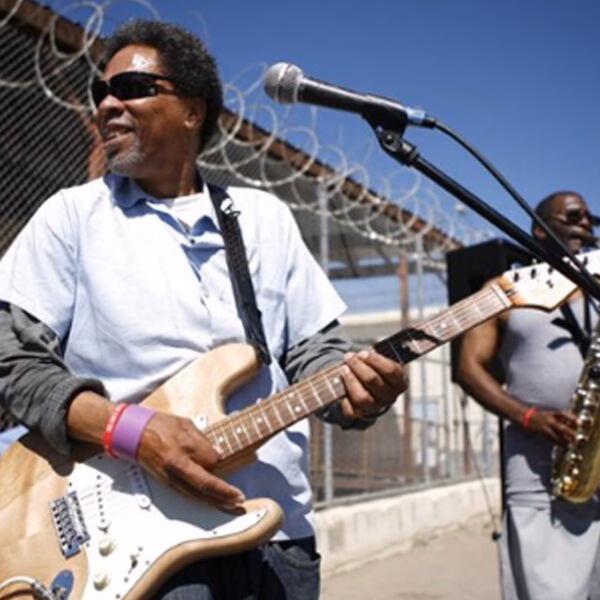 A man plays an electric guitar while another musician plays a saxophone in front of a prison fence with barbed wire.