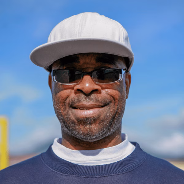 A close-up portrait of a smiling man with a grey beard, wearing a white baseball cap and dark sunglasses, against a bright blue sky.