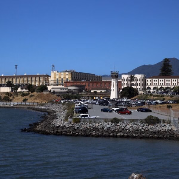 A coastal view of the Alcatraz Island prison complex, featuring a prominent white lighthouse, multi-story buildings, and a parking lot, with a mountain in the background under a clear blue sky.