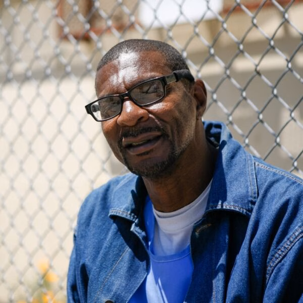A close-up portrait of a smiling Black man with glasses, wearing a blue denim jacket over a blue shirt, in front of a chain-link fence.