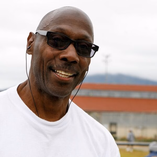 A smiling man with a shaved head, wearing sunglasses and a white t-shirt, with earphones in his ears.