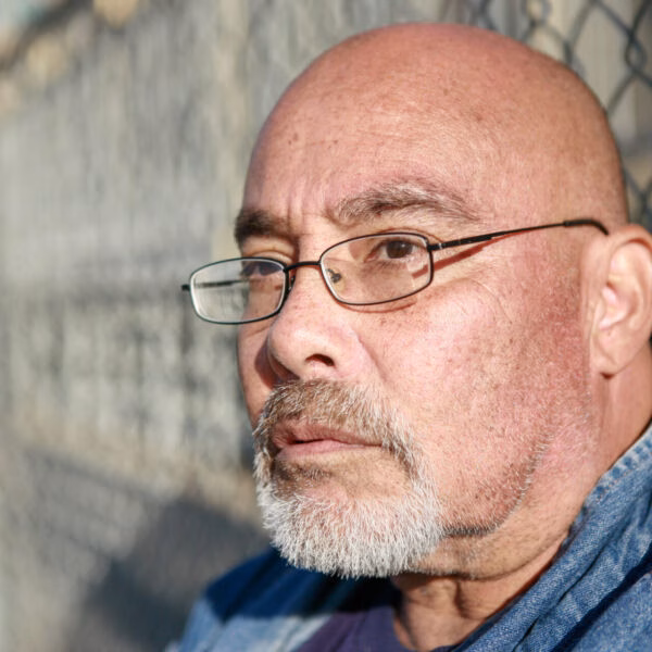A close-up portrait of a bald man with glasses and a grey beard, wearing a denim shirt, looking thoughtfully to the side against a chain-link fence.