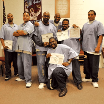 Seven men in light blue prison uniforms holding certificates, posing for a group photo in a church setting.