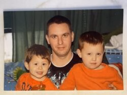 A man with short dark hair wearing a black t-shirt sits between two young boys in orange shirts.