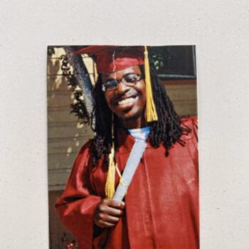 A smiling man with glasses and dreadlocks in a red graduation cap and gown, holding a diploma.