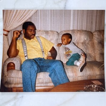 A man in a yellow shirt and suspenders sits on a couch with a young child laughing beside him.