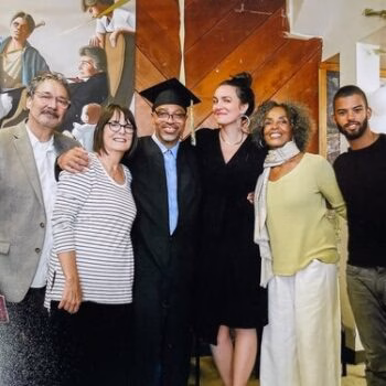 Six people pose for a group photo in front of a large mural. A man in a graduation cap stands in the center, flanked by five others in a casual indoor setting.