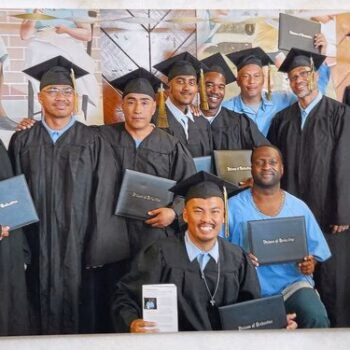 A group of nine graduates in black caps and gowns holding blue diplomas, posing for a group photo.