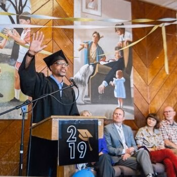 A man in a graduation cap and gown stands at a podium marked '2019', gesturing while speaking into a microphone. Behind him, a mural depicts historical figures, and a group of people sits in the audience.