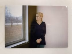 A woman with blonde hair in a dark top stands by a window, looking out at a winter landscape.