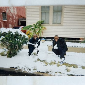 Two men in dark jackets crouch in a snow-covered yard, smiling as they pose with a small snowman.