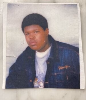 Portrait of a young man with dark hair wearing a blue denim jacket with 'Pamphile' embroidered on the left chest over a white graphic t-shirt.