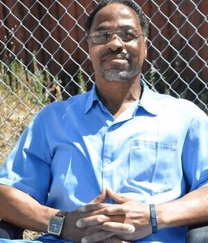A man with glasses and a beard, wearing a blue shirt, sits in front of a chain-link fence.