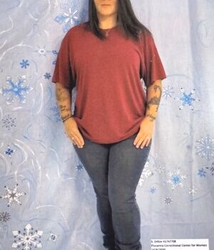 Shebri2 A woman with long dark hair and tattoos on both arms stands in front of a blue patterned backdrop.