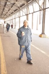 Thomas stands in a covered walkway, wearing a grey jacket and pants with 'MIZOUI' and 'SCR' text, eating a sandwich.