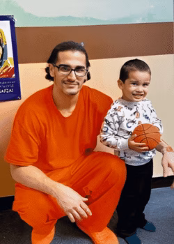 Daniel, 31, in an orange jumpsuit, smiles while kneeling next to a young boy holding a basketball.