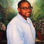 A man with glasses and a light blue shirt stands in front of a tropical painting.