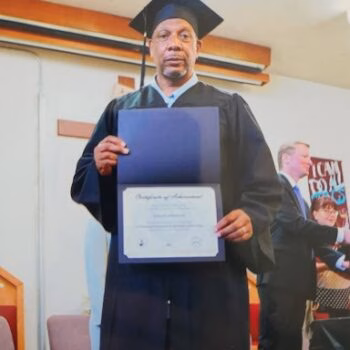 A man in a black graduation cap and gown holds a certificate of achievement.