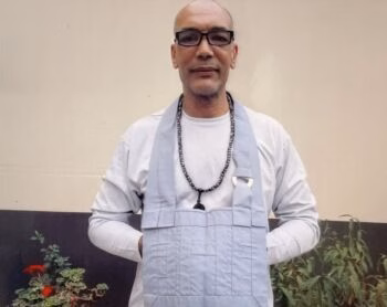 Mark, 59, a bald man with glasses and a beard, wearing a light blue apron over a white shirt and a beaded necklace, stands against a plain wall with plants.