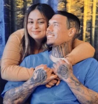 A man and woman embrace in a forest setting. The man has extensive tattoos on his neck and arms, including a '100' on his hand.