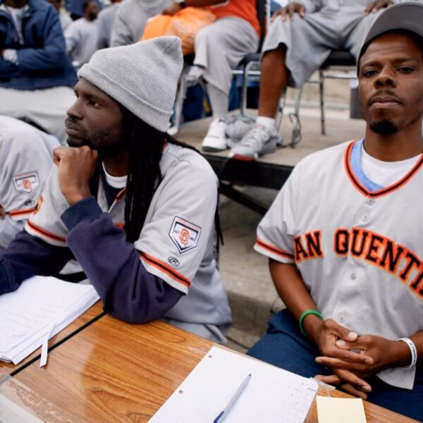 Three men in San Quentin baseball jerseys sit at a table with papers and pens, appearing to be in a classroom or workshop setting.