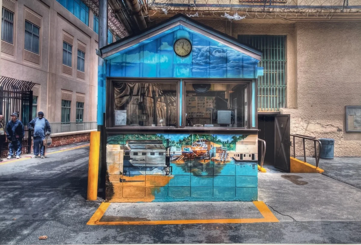 An outdoor photograph of a small security guard shack painted with a vibrant blue sky and a waterfront mural, situated between urban buildings with two people standing on the left.