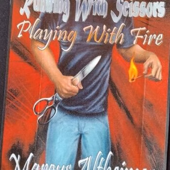 Marcus 2 A man in a blue shirt holds a knife and scissors in one hand and lights a match with the other against a red background.