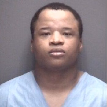 Mugshot of a Black man with short hair wearing a light blue V-neck shirt against a grey background.