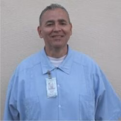 Edwin, a smiling man in a light blue work shirt with an ID badge, stands against a plain wall.