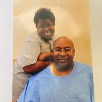 A man in blue medical scrubs and glasses sits in front of a smiling woman in a grey shirt, both posing for a photo.