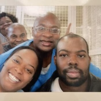 Five Black individuals, including a man in a blue shirt and glasses, pose for a selfie indoors.