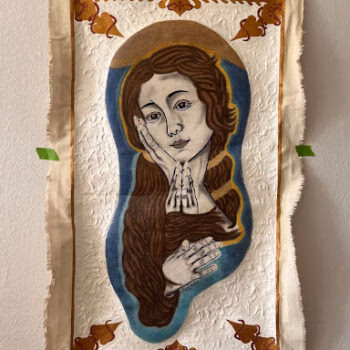 A religious icon of the Virgin Mary with a halo, her hand to her cheek, set within a blue oval on a textured white fabric background with decorative golden motifs.