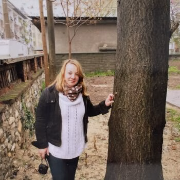 Aharon003 (4) A woman with blonde hair, wearing a white sweater and black jacket, stands next to a large tree trunk in an outdoor area.
