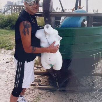 Aharon007 (1) A man named Aharon007 holds a white torso mannequin in front of a green boat on a sandy beach.