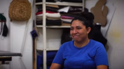 Blanca, a woman with dark hair in a bun, wearing a blue t-shirt, smiles while being interviewed in a workshop.