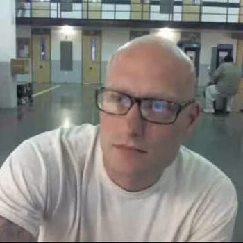 A bald man with glasses and a white t-shirt, identified as Brian 2, looks at the camera in a large indoor space.