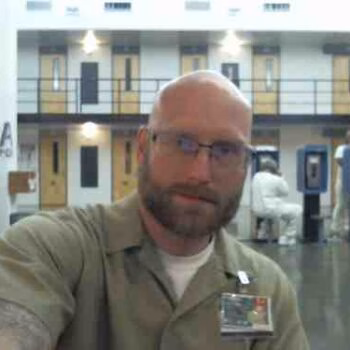 A bald man with glasses and a beard wearing a tan uniform shirt with a name tag, standing in a prison cell block.
