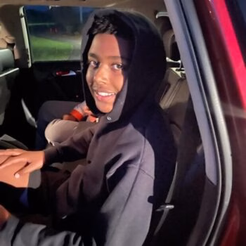 A young man with a dark hooded sweatshirt sits in the back of a car at night, smiling at the camera.