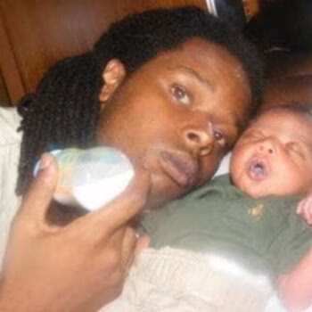 A man with dreadlocks holds a baby bottle next to a sleeping newborn baby.