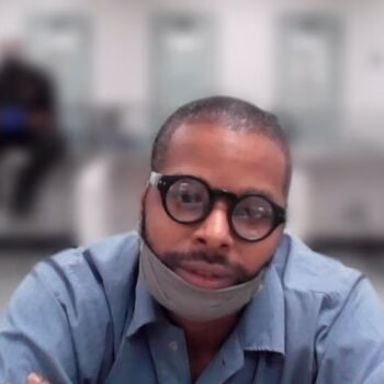 A man with glasses and a pulled-down mask looks at the camera in a blurred indoor setting.