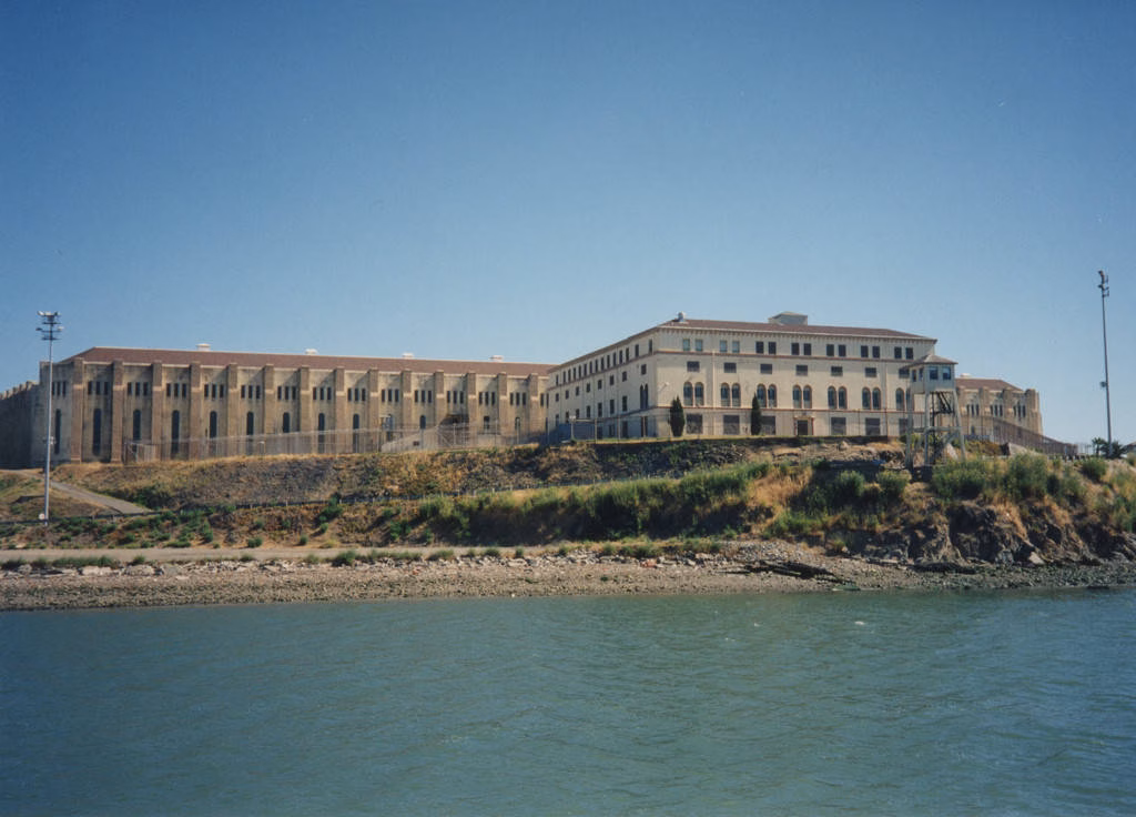 A large, multi-winged institutional building complex situated on a grassy hill overlooking a body of water under a clear blue sky.