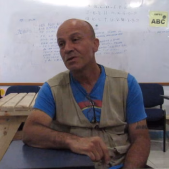 A man in a blue shirt and beige vest sits at a desk in a classroom, with a whiteboard behind him.