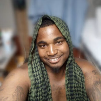 A portrait of Kunta, a man with a goatee and tattoos, smiling while wearing a green patterned cloth over his head.