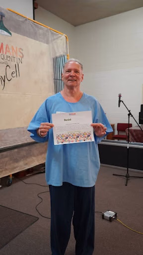 Daniel N. holds a certificate with his name and a colorful illustration, standing in front of a banner for the Mans Quintin Family Cell.